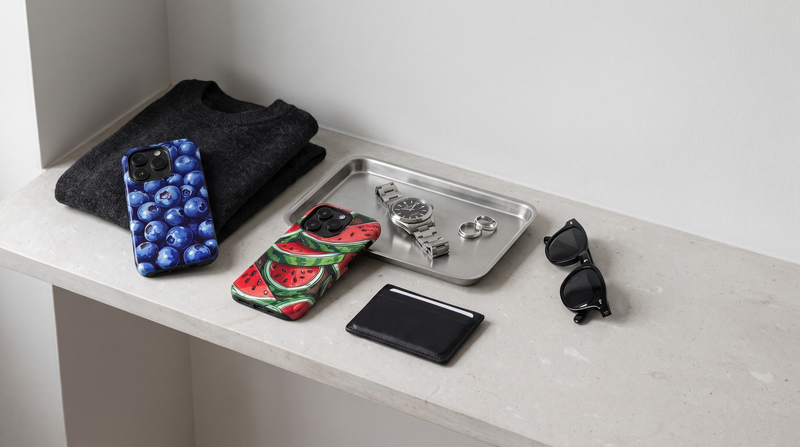 Blueberry and Watermelon Art iPhone tough cases on a limestone shelf beside a watch, sunglasses and a folded knit